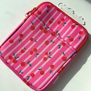 May include: Pink and white striped tablet sleeve with a pattern of red cherries and green leaves. The sleeve has a red zipper and trim. The text "@Kelleandco" is visible in the upper right corner.