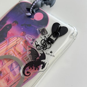 May include: A clear phone case featuring a fantasy-themed graphic with a couple, a moon, and framed pictures. It includes a black heart-shaped charm, a small book charm with an S, a sword charm, and a black dragon charm attached by a black clasp.