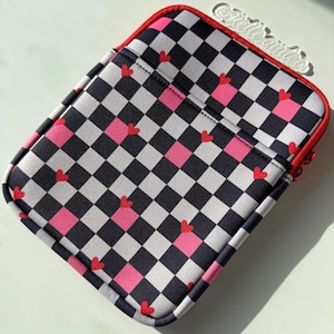 May include: A padded tablet sleeve with a red zipper and trim. The sleeve features a black, white, and pink checkered pattern with small red hearts scattered across the surface. The sleeve has a pocket on the front.