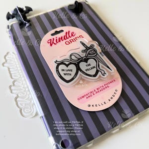 May include: A clear Kindle grip with a pink backing card. The grip features two heart-shaped designs with the text "IN LOVE WITH" and "THE VILLAIN", and a sword. The background is a purple and black striped pattern.