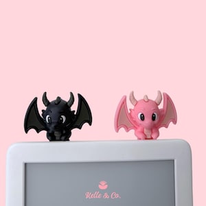May include: Two small dragon-shaped figures, one black and one pink, are perched on the top edge of a white rectangular frame. The frame features the text Kelle & Co. in pink lettering. Each dragon has horns, wings, and large eyes.