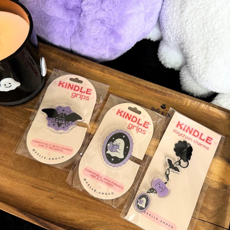 May include: Three Kindle accessories on a wooden tray. Two are Kindle grips with a bat and ghost design, and the third is a keychain with a bat and ghost charm. The grips and keychain are packaged on pink and white cards.