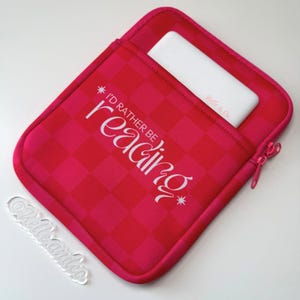 May include: A pink and red checkered sleeve with the text "I'd rather be reading" in white. A white rectangular object is inside the sleeve. A clear acrylic tag with the text "Belle & Co" is on the bottom left.