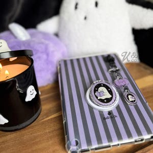 May include: A Halloween-themed collection featuring a black candle with ghost decals, a purple striped tablet case, and a ghost-themed phone grip and keychain. The items are arranged on a wooden surface.