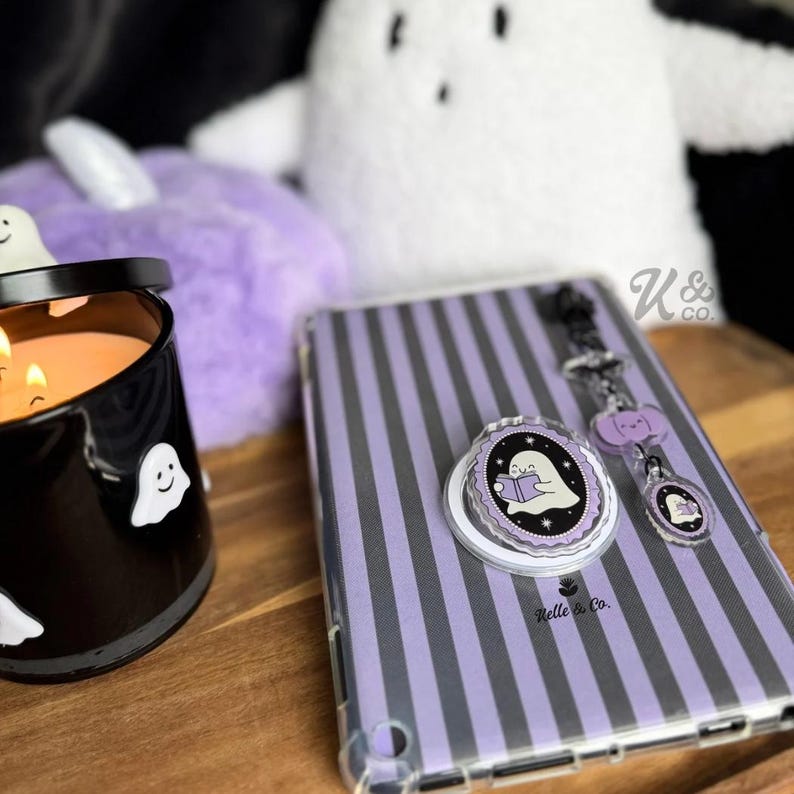 May include: A Halloween-themed collection featuring a black candle with ghost decals, a purple striped tablet case, and a ghost-themed phone grip and keychain. The items are arranged on a wooden surface.