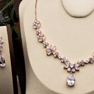 May include: A rose gold bridal necklace and earrings set with a teardrop pendant and floral design. The jewelry is adorned with clear cubic zirconia stones.