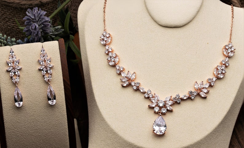 May include: A rose gold bridal necklace and earrings set with a teardrop pendant and floral design. The jewelry is adorned with clear cubic zirconia stones.