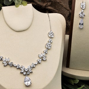 May include: A silver necklace and earrings set with a teardrop pendant and floral accents. The necklace features a delicate chain with a large, pear-shaped crystal in the center.