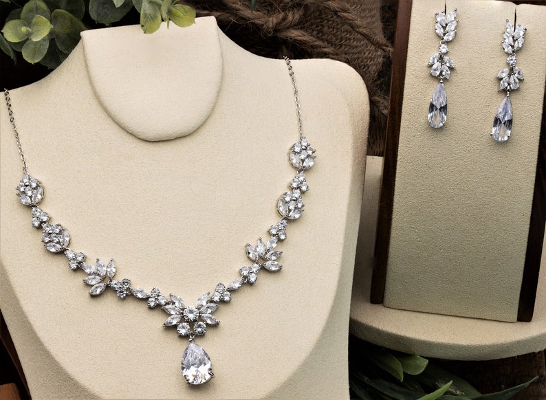 May include: A silver necklace and earrings set with a teardrop pendant and floral accents. The necklace features a delicate chain with a large, pear-shaped crystal in the center.