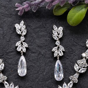 May include: A silver necklace and earrings set with a teardrop-shaped pendant and a delicate floral design. The jewelry is made with clear cubic zirconia stones.