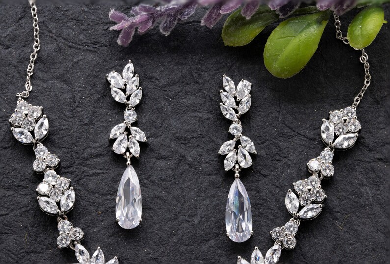 May include: A silver necklace and earrings set with a teardrop-shaped pendant and a delicate floral design. The jewelry is made with clear cubic zirconia stones.