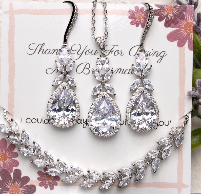 May include: A silver bridal jewelry set with a teardrop pendant necklace and matching earrings. The necklace and earrings feature a pear-shaped crystal surrounded by smaller crystals in a floral design.