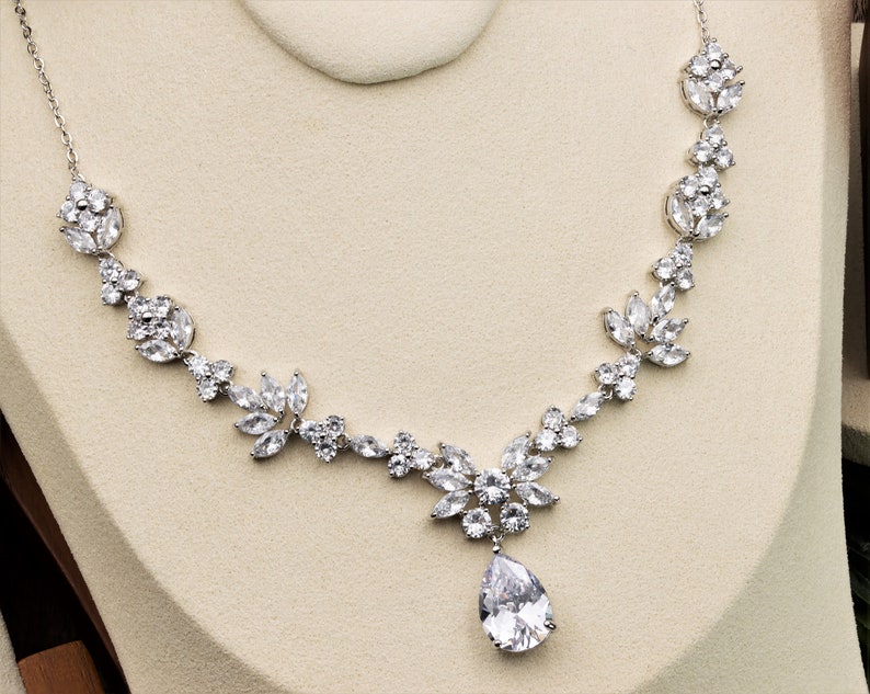 May include: A silver necklace with a teardrop-shaped pendant and a floral design. The necklace is made of clear cubic zirconia stones.