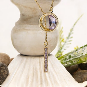 Può includere: Una collana con catena dorata con un ciondolo di cristallo viola. Il ciondolo è un cerchio con un cerchio più piccolo all'interno, e il cristallo è posto al centro. La collana ha anche una lunga e sottile barra di cristallo viola che pende dal basso.