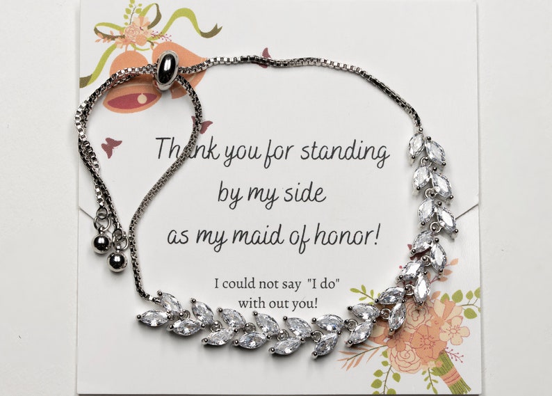 May include: A silver bracelet with a delicate chain and a charm featuring a cluster of clear rhinestones. The bracelet has a pull-string closure. The card reads "Thank you for standing by my side as my maid of honor! I could not say "I do" with out you!"