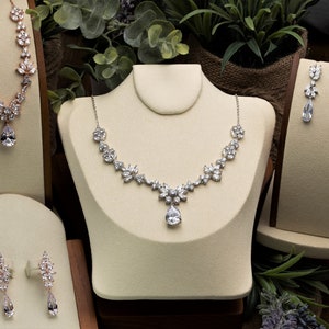May include: Three sets of jewelry displayed on velvet stands. The first set is a rose gold necklace with a teardrop pendant. The second set is a silver necklace with a teardrop pendant and a matching pair of earrings. The third set is a pair of silver earrings with teardrop pendants.