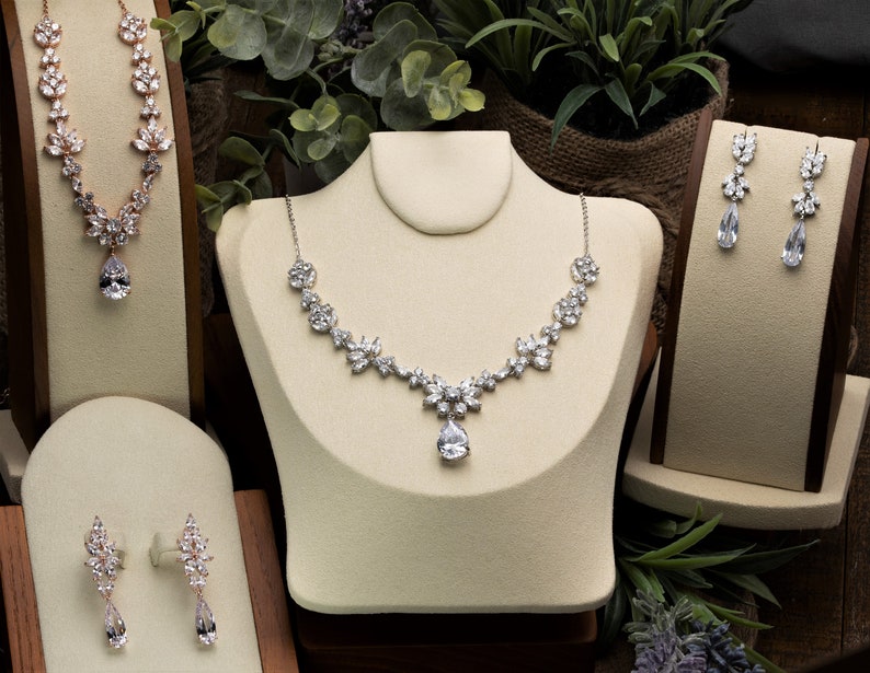 May include: Three sets of jewelry displayed on velvet stands. The first set is a rose gold necklace with a teardrop pendant. The second set is a silver necklace with a teardrop pendant and a matching pair of earrings. The third set is a pair of silver earrings with teardrop pendants.