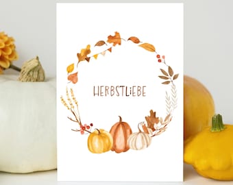 May include: A white greeting card featuring a watercolor illustration of an autumn wreath made of orange, yellow, and brown leaves, berries, and three pumpkins at the bottom. The text HERBSTLIEBE is printed in brown in the center of the wreath.
