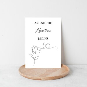 May include: A white card with black text that reads "And so the adventure begins" with a simple line drawing of a rose and two rings connected by a heart.
