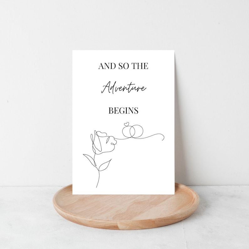 May include: A white card with black text that reads "And so the adventure begins" with a simple line drawing of a rose and two rings connected by a heart.