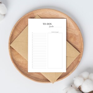 May include: A white printable to-do list with a section for notes. The to-do list has a title that says "To-Dos" and a line for writing in the week. The notes section has the title "Anmerkungen". The printable is on a wooden plate with a brown envelope.