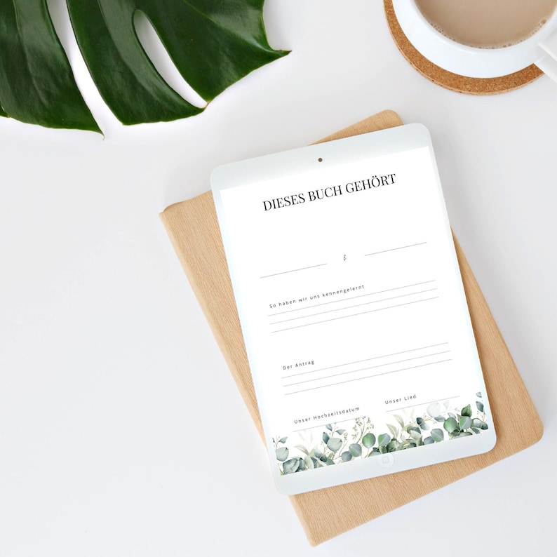 Pode incluir: Um tablet branco com uma capa de madeira marrom. A tela do tablet exibe um livro de visitas de casamento imprim&iacute;vel com o texto "Dieses Buch geh&ouml;rt" e um design floral na parte inferior.