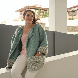 May include: A teal blue knitted cardigan with a shawl collar and pockets. The cardigan has a contrasting white and brown knit trim on the sleeves and pockets.