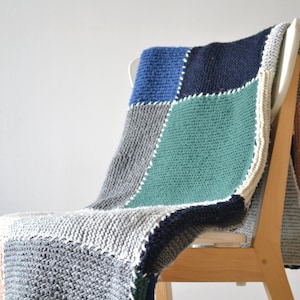 Puede incluir: Una manta de patchwork de punto en tonos grises, azules, verdes y rosas, drapeada sobre una silla de madera.