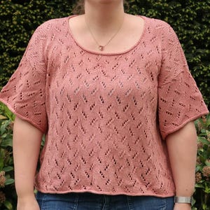 Puede incluir: Una blusa de punto rosa polvoriento de manga corta, con cuello redondo y un delicado diseño calado. La blusa se combina con vaqueros azules. El estilo general es informal y cómodo.