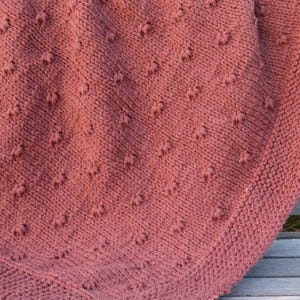 May include: A dusty rose-colored knitted blanket with a textured pattern of raised stitches. The blanket is draped over a wooden bench, showcasing its soft texture and detailed design. The blanket's edges have a ribbed border.