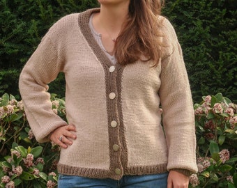 May include: A woman wearing a light brown cardigan with a brown trim and a v-neck. She is wearing blue jeans and has brown hair and is wearing sunglasses.