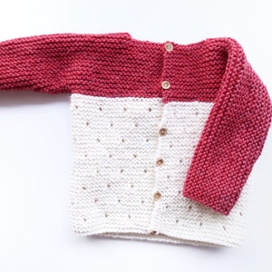Peut inclure: Un cardigan en tricot pour bébé avec des manches longues. Le cardigan est blanc avec une bande rouge en haut et en bas. Il a une fermeture boutonnée sur le devant.