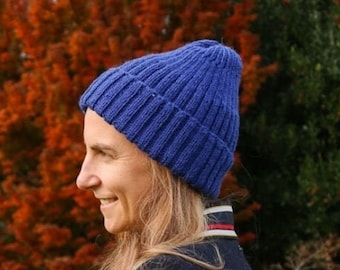 May include: A blue knitted hat with a ribbed texture. The hat is worn by a person with long blonde hair, standing in front of a background of fall foliage.
