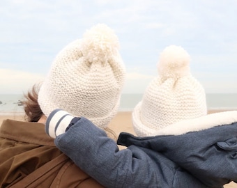 Könnte beinhalten: Eine Frau in einer braunen Jacke hält ein Kind, das eine blaue Jacke und eine weiße Strickmütze mit Bommel trägt. Das Kind trägt eine weiße Strickmütze mit Bommel. Das Bild trägt die Bildunterschrift "Druckbarer PDF-Strickmuster".