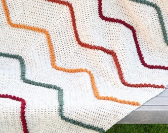 May include: A white crocheted blanket with a chevron pattern. The blanket has a border of colorful yarn pom-poms in shades of red, orange, green, and brown.