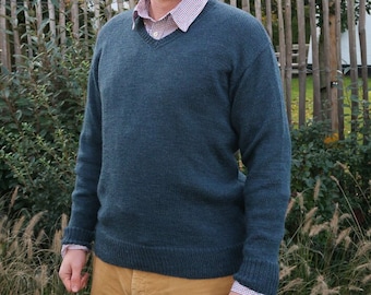 May include: A man wearing a dark blue sweater over a light blue and white checkered shirt. He is wearing khaki pants and is standing in front of a wooden fence.