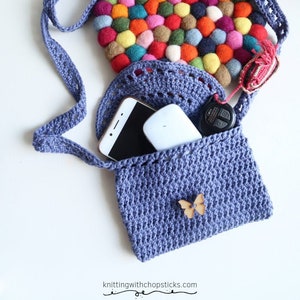 May include: A crocheted blue crossbody bag with a colorful felt circle inside. The bag has a small wooden butterfly charm and is open to reveal a phone and keys.
