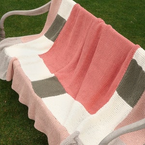 May include: A knitted throw blanket with a patchwork design in shades of pink, white, and gray. The blanket is folded and draped over a wooden bench.