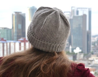 May include: A gray knit beanie is worn by a person looking out at a city skyline. The beanie is ribbed at the brim and has a textured, vertical pattern. The person is wearing a maroon top with a textured pattern.