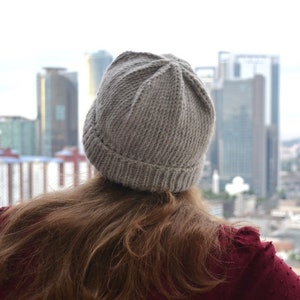 May include: A gray knit beanie is worn by a person looking out at a city skyline. The beanie is ribbed at the brim and has a textured, vertical pattern. The person is wearing a maroon top with a textured pattern.