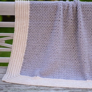 May include: A crocheted blanket with a light gray center and a cream colored border. The blanket is draped over a wooden bench.