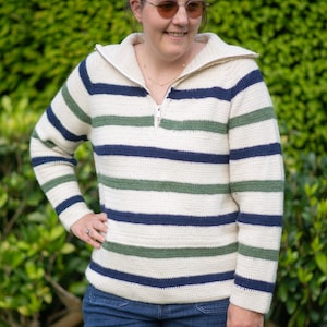 Puede incluir: Un suéter de crochet a rayas con una cremallera en la parte delantera. El suéter tiene rayas blancas, verdes y azules. La persona que lleva el suéter está de pie frente a un arbusto verde.