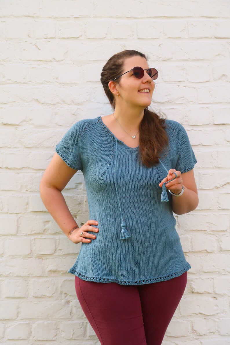 Pu&ograve; includere: Un top in maglia color teal a maniche corte, con scollo a V e laccetto con nappe. Il top presenta un orlo e bordi delle maniche decorativi. La modella indossa pantaloni bordeaux. Lo sfondo &egrave; un muro di mattoni bianchi.
