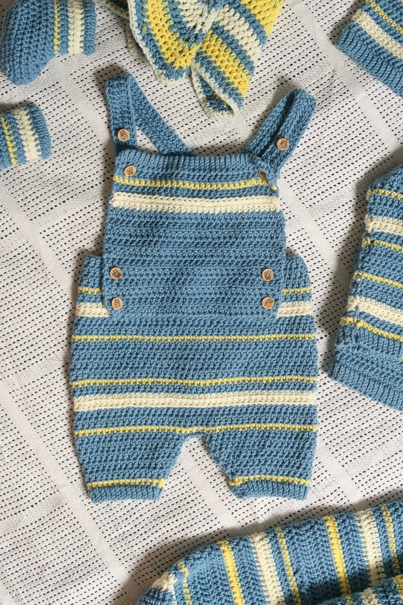 May include: A blue and white striped crocheted baby outfit with yellow accents. The outfit includes a one-piece jumpsuit with straps and a matching hat.