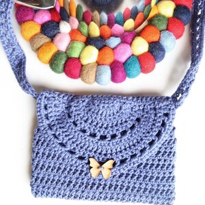 May include: A small, lavender crocheted bag with a wooden butterfly embellishment. The bag has a long strap and is decorated with a colorful circle of felt balls.