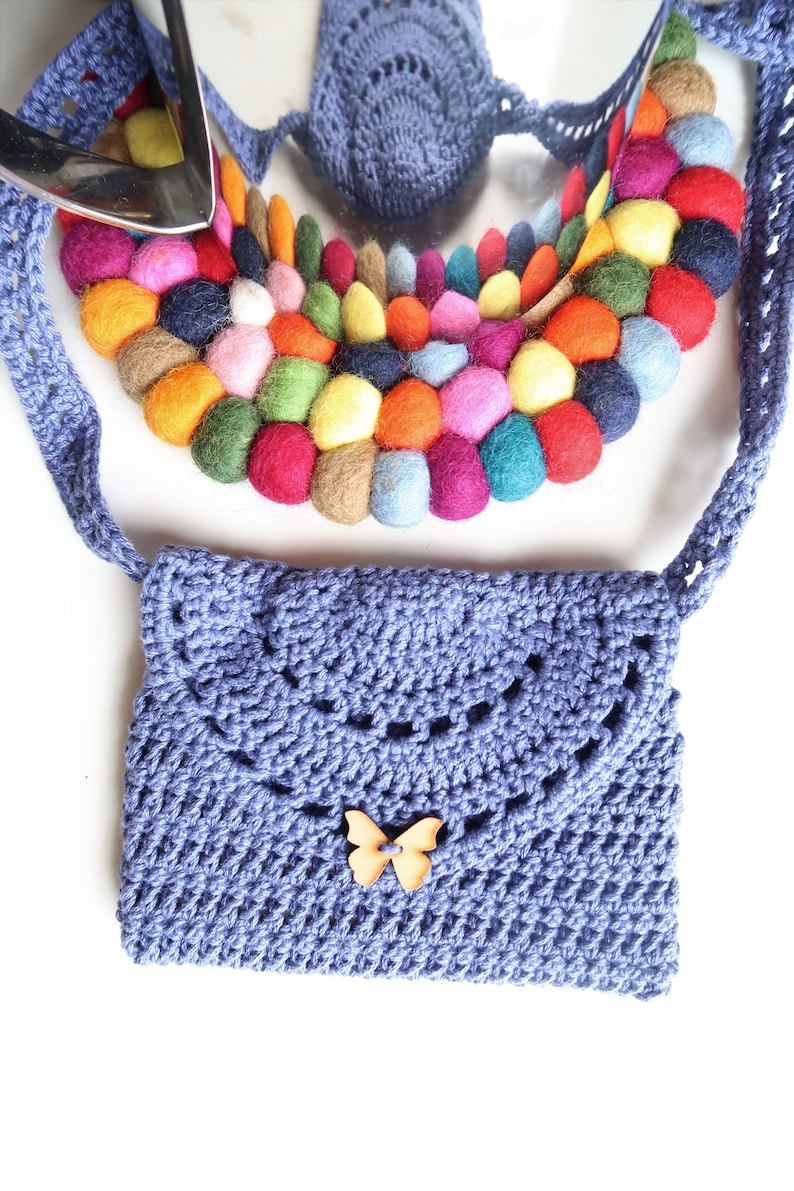 May include: A small, lavender crocheted bag with a wooden butterfly embellishment. The bag has a long strap and is decorated with a colorful circle of felt balls.