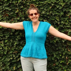 May include: A woman wearing a turquoise blue knitted top with a V-neck and short sleeves. The top has a ruffled hem and is worn with light gray pants.