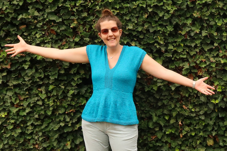May include: A woman wearing a turquoise blue knitted top with a V-neck and short sleeves. The top has a ruffled hem and is worn with light gray pants.