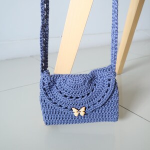 May include: A small, crocheted purse with a butterfly-shaped button. The purse is a light blue color and has a long strap.