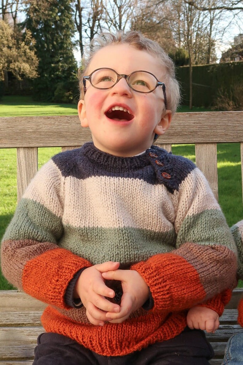 Op de afbeelding: Een jong kind draagt een gestreepte gebreide trui met oranje, groene, beige en blauwe strepen. De trui heeft een knoopsluiting op de schouder. Het kind zit op een houten bankje en kijkt omhoog.